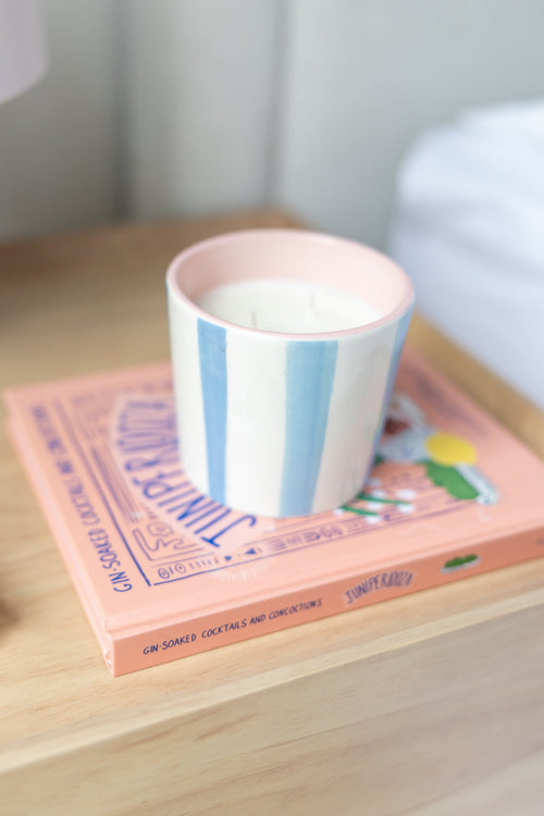 A pastel pink and white striped candle jar sits on top of a pink book on a light wood surface, with a softly blurred home interior in the background.