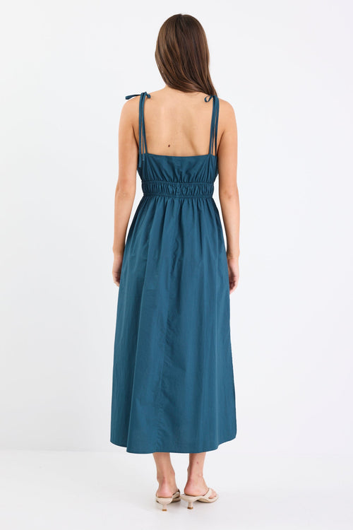 Woman wearing a petrol blue strappy cotton midi dress with a tie detail at the bust, gathered waist, and side splits.