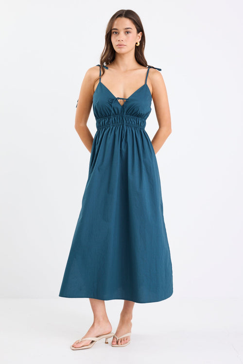 Woman wearing a petrol blue strappy cotton midi dress with a tie detail at the bust, gathered waist, and side splits.