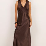 A woman modeling a sleeveless, wrap-style midi dress in a dark brown satin-like fabric, standing against a plain off-white studio background.