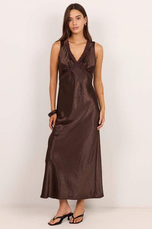 A woman modeling a sleeveless, wrap-style midi dress in a dark brown satin-like fabric, standing against a plain off-white studio background.