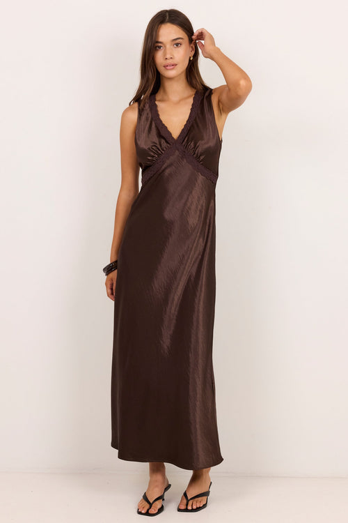 A woman modeling a sleeveless, dark brown satin maxi dress with a V-neckline and draped waist details, standing against a plain white studio background.