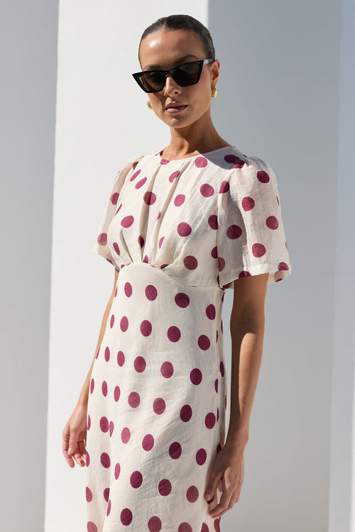 A woman modeling a cream-colored midi dress with dark red polka dots, styled with black sunglasses, standing in a bright white architectural setting with strong shadows.