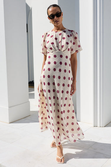 A woman wearing a cream midi dress with red polka dots and sunglasses, walking outdoors on a white tiled patio in front of a white building with columns.
