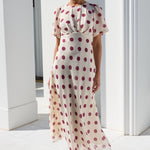 A woman wearing a cream midi dress with red polka dots and sunglasses, walking outdoors on a white tiled patio in front of a white building with columns.
