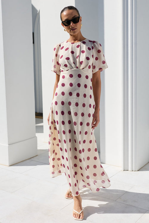 A woman wearing a cream midi dress with red polka dots and sunglasses, walking outdoors on a white tiled patio in front of a white building with columns.