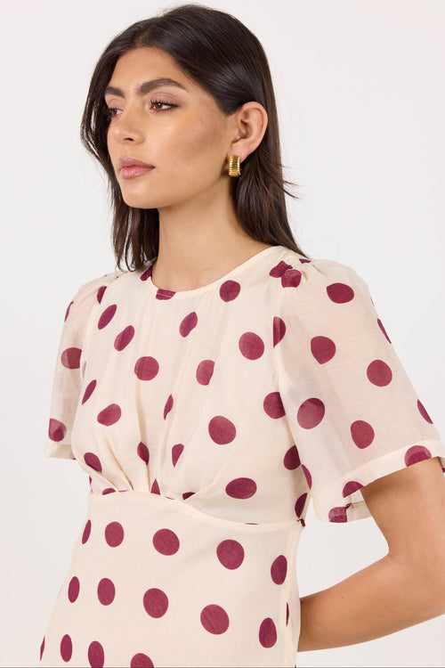 A woman wearing a short-sleeve cream-colored dress with large dark red polka dots, photographed against a plain light background.