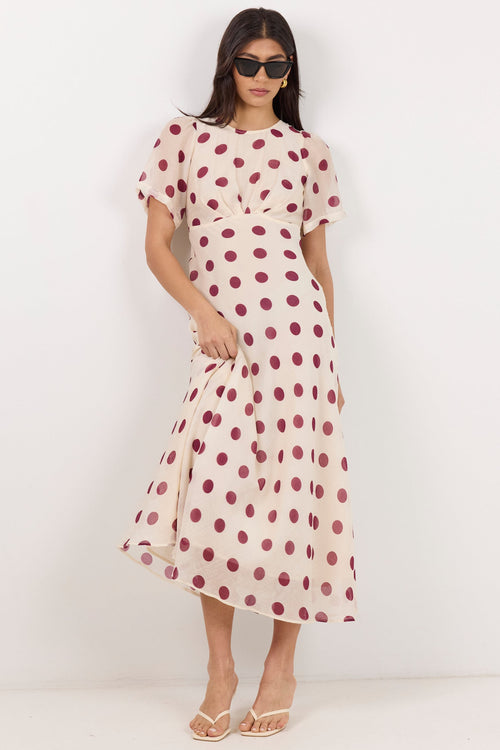 A woman modeling a short-sleeve midi dress with a cream base and dark red polka dots, paired with beige flat sandals and sunglasses, set against a plain white studio background.