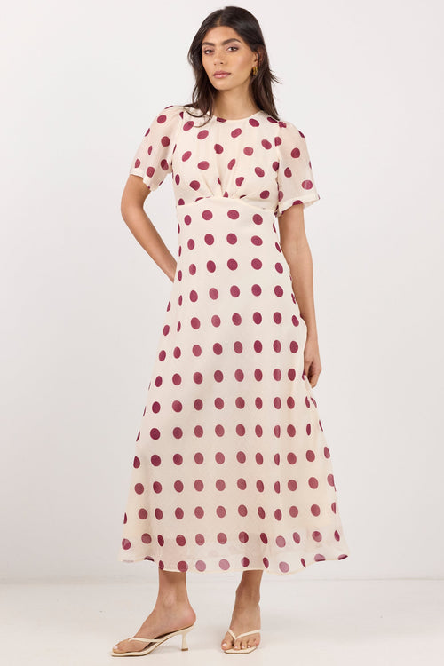 A woman modeling a short-sleeve midi dress with a pink and white polka dot pattern, standing against a plain white studio background.