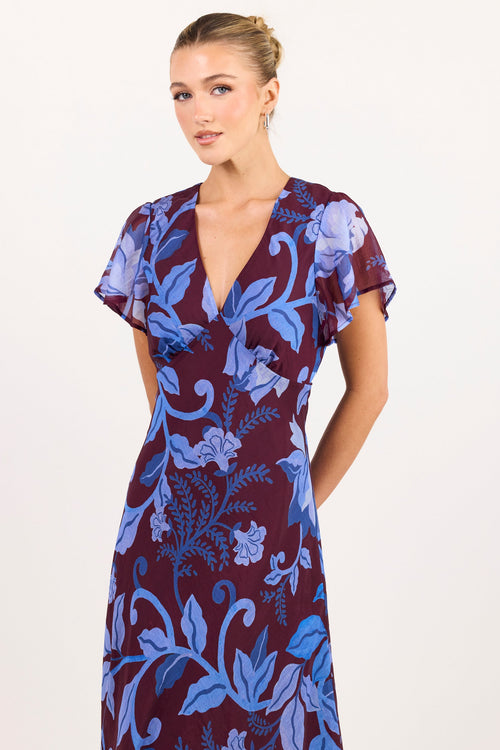 A woman modeling a short-sleeve wrap dress with a deep V-neckline, featuring a blue floral and vine pattern on a deep burgundy background, set against a plain white studio backdrop.