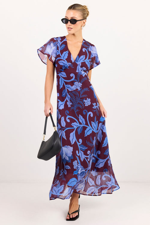 A woman modeling a long, flowy wrap-style dress with flutter sleeves and a blue floral print on a deep purple background, styled with black sunglasses, a black handbag, and black sandals against a plain light studio backdrop.