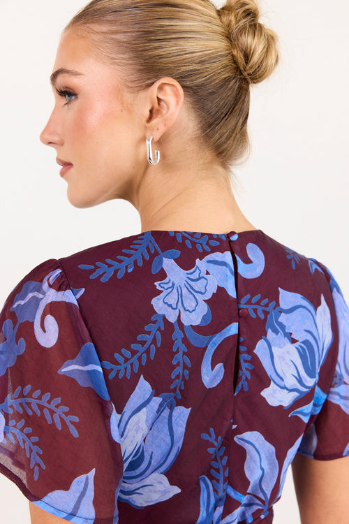 A woman shown from the back wearing a sheer short-sleeve top with a blue floral print on a deep burgundy background. She is wearing silver hoop earrings against a plain off-white background.