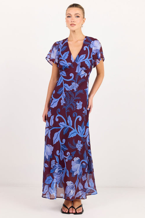 A woman modeling a long wrap-style maxi dress with a blue and purple floral print, standing against a plain white studio background.