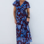 A woman modeling a short-sleeve wrap maxi dress with a blue and purple floral pattern, standing against a minimalist white wall with a circular arch.