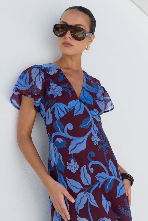 A woman wearing a dark burgundy maxi dress with a blue floral print, ruffled short sleeves, and a V-neckline, posing against a white arched wall.