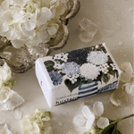 A boxed fragrance product labeled 'huxley' and 'CAROLINE HALL' lies on a glossy white surface scattered with white hydrangea petals and flowers. The box features a floral print in white, blue, and green, with a silver bowl of white hydrangeas in the upper left.