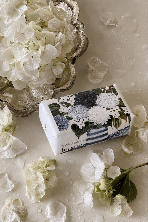 A boxed fragrance product labeled 'huxley' and 'CAROLINE HALL' lies on a glossy white surface scattered with white hydrangea petals and flowers. The box features a floral print in white, blue, and green, with a silver bowl of white hydrangeas in the upper left.