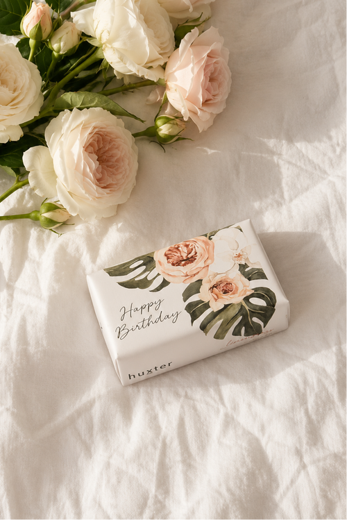 A wrapped gift box with floral artwork and the words 'Happy Birthday' lies on white fabric next to a bouquet of pale blush and cream roses with green leaves.