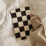 A black and cream checkerboard notebook with gold 'D'KIS' lettering, held by a person wearing a beige knit sweater over rumpled white bedding.