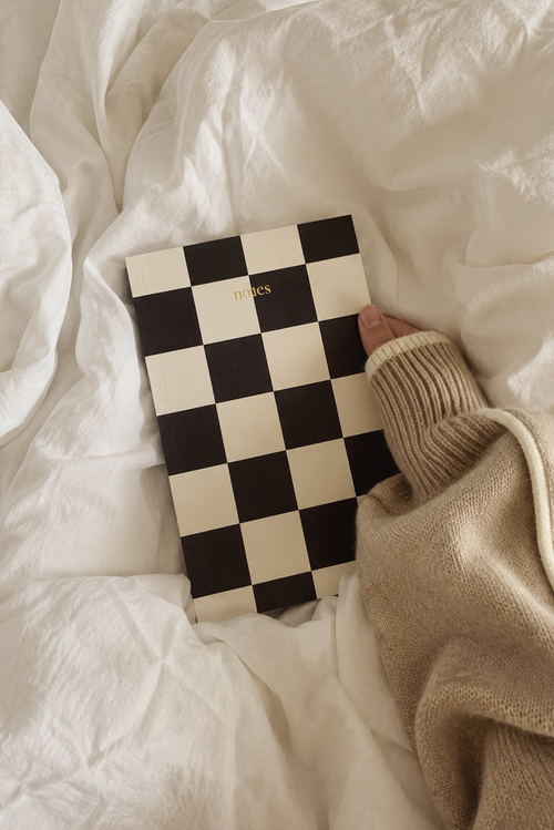 A black and cream checkerboard notebook with gold 'D'KIS' lettering, held by a person wearing a beige knit sweater over rumpled white bedding.