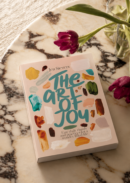 A book titled "The Art of Joy" by Jen Sievers lies on a white marble tray, styled with two purple tulips in a glass vase on a textured cream fabric background.