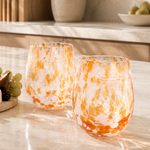 Two stemless wine glasses with an orange and white marbled pattern, set on a light marble kitchen counter next to a charcuterie board with cheese, grapes, and crackers.