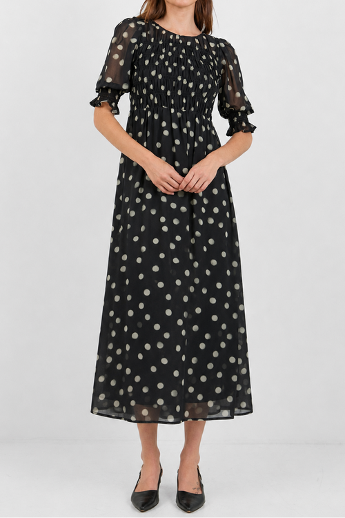 A woman modeling a black midi dress with white polka dots, featuring sheer puff sleeves and a gathered waist, set against a plain light gray background.