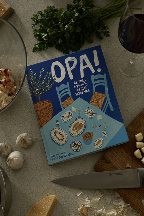 A cookbook titled 'OPA!' lies on a kitchen counter surrounded by food prep items including a knife, garlic cloves, parsley, a glass of red wine, butter, and cheese.