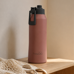 A dusty rose pink Fresko water bottle with a dark gray lid and carrying loop, standing on a light wood bench next to a beige woven bag.