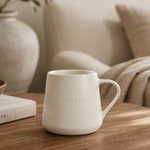 A matte off-white ceramic mug with embossed text reading 'Hug by A Man' sits on a round wooden table in a cozy living room setting.