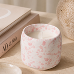 A white ceramic candle jar with a pink speckled pattern, styled on a beige surface next to a small dish, books, and a textured vase.