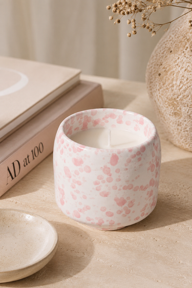 A white ceramic candle jar with a pink speckled pattern, styled on a beige surface next to a small dish, books, and a textured vase.
