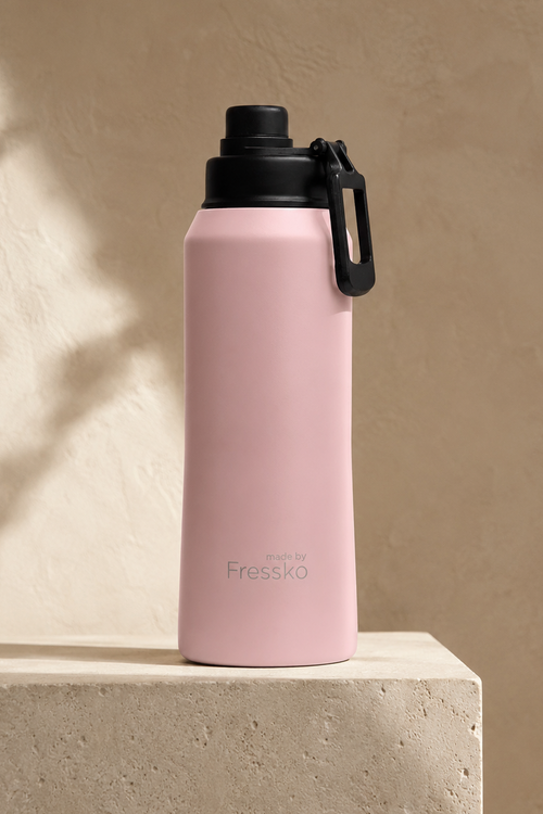 A pink insulated water bottle with a black lid and carrying loop, standing on a textured beige stone pedestal against a beige wall with soft leaf shadows.