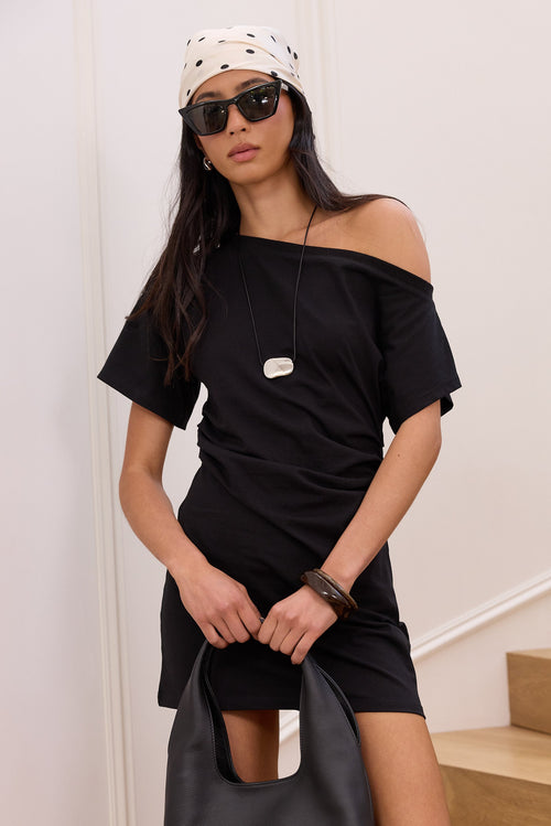 A woman models a black off-the-shoulder short-sleeve mini dress, styled with a black leather hobo bag, black sunglasses, a black and white polka dot bandana, and a silver pendant necklace, standing in an indoor setting with white walls and wooden stairs.