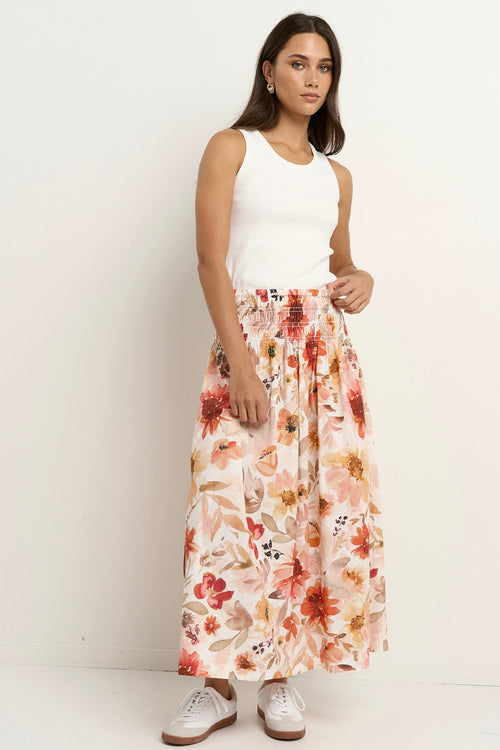 Woman wears a floral maxi skirt with a tiered silhouette, elastic waist, and white ribbed tank top with sneakers.
