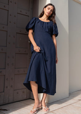 Woman wearing a navy puff-sleeve maxi dress with a gathered scoop neckline and bias-cut silhouette, styled with white sandals in a light architectural setting.







