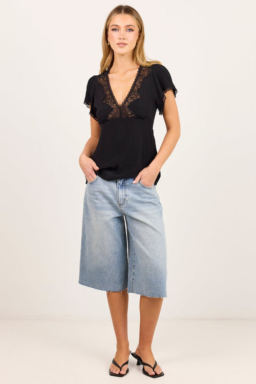 A woman modeling a black short-sleeve top with a lace V-neckline, paired with light-wash blue denim culottes and black sandals, standing against a plain off-white studio background.