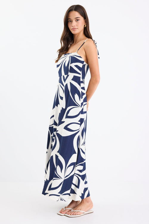 Woman wearing a navy and white printed maxi dress with tie straps, shirred back, and a flowing full-length silhouette.