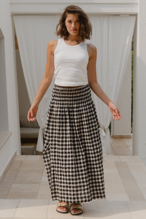 Woman wearing a white sleeveless top and black and white checkered skirt standing in a sunlit outdoor setting.