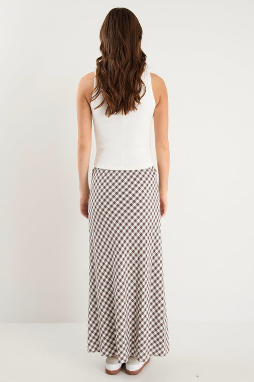 Woman wearing a brown and white gingham print maxi skirt with a tie waist and bias cut, paired with a fitted white tank and white sneakers.