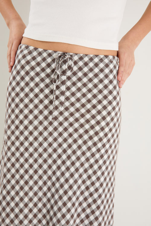Woman wearing a brown and white gingham print maxi skirt with a tie waist and bias cut, paired with a fitted white tank and white sneakers.