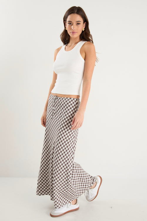 Woman wearing a brown and white gingham print maxi skirt with a tie waist and bias cut, paired with a fitted white tank and white sneakers.