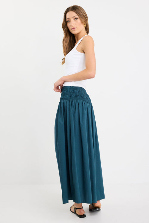 Woman wearing a petrol blue cotton maxi skirt with wide shirred waistband, styled with a white tank and black sandals.