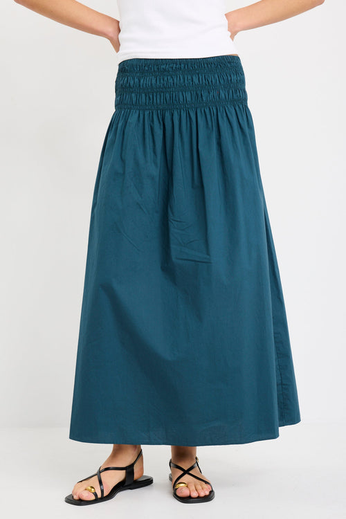 Woman wearing a petrol blue cotton maxi skirt with wide shirred waistband, styled with a white tank and black sandals.