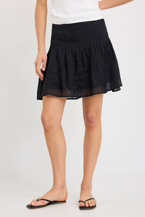 Woman wearing a black mini skirt with pintuck waist detail and soft flared shape, styled with a white tank and black sandals.