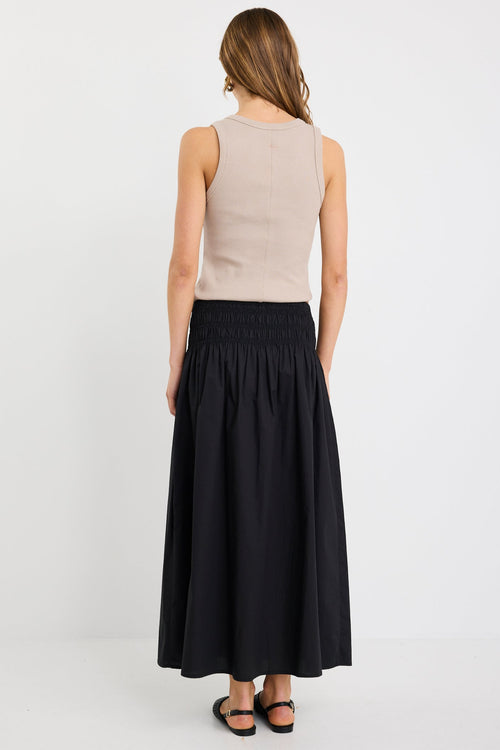 Woman wearing a black cotton maxi skirt with shirred waistband, styled with a taupe ribbed tank and black woven flats.