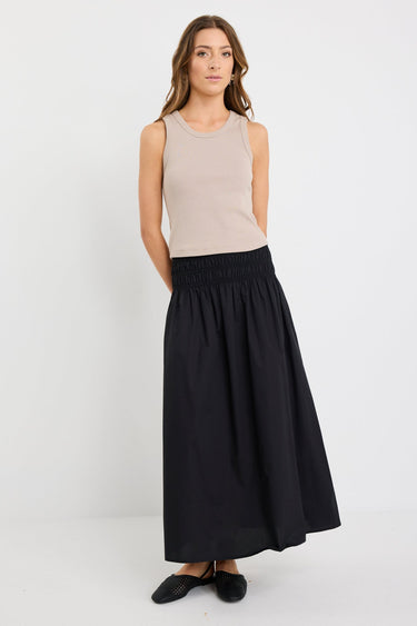 Woman wearing a black cotton maxi skirt with shirred waistband, styled with a taupe ribbed tank and black woven flats.
