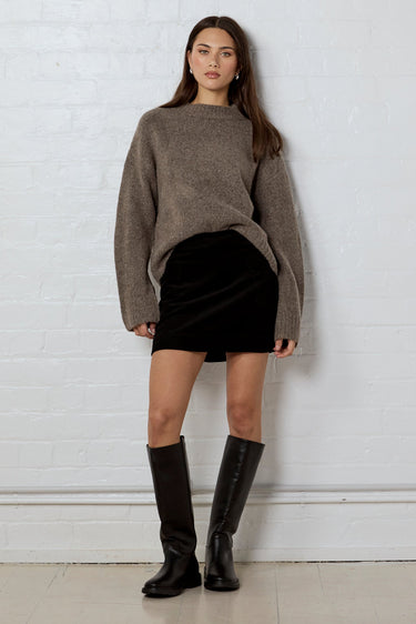 Woman wearing a brown sweater, black skirt, and knee-high boots against a white brick wall.