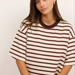 A woman wearing a brown and cream striped short-sleeve t-shirt with a solid brown crew neck, paired with light blue jeans, seated against a plain off-white background.