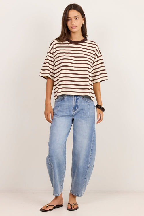 A woman modeling a brown and white horizontally striped short-sleeve t-shirt with light-wash blue jeans and brown sandals, standing against a plain off-white studio background.